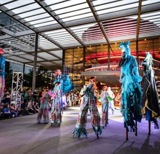Performers on stilts in colorful costumes entertain a crowd outside a modern, glass-fronted building at night.
