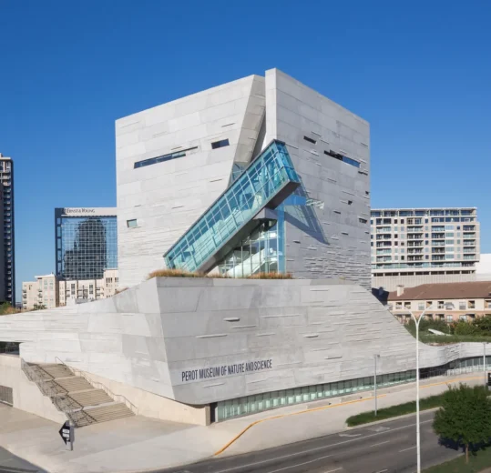 Modern building with angular, white stone architecture and glass accents, surrounded by city skyscrapers under a clear blue sky.