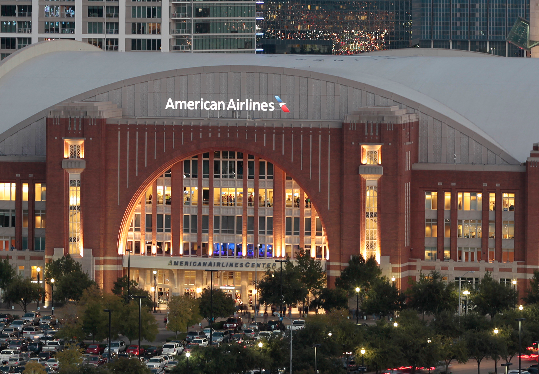 The American Airlines Center in Dallas, Texas, is shown at dusk with lights on and a full parking lot in front.