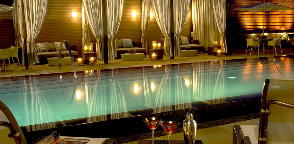 Outdoor pool at night with cabanas, lounge seating, candles, and two cocktails on a table in the foreground, creating a relaxing, elegant atmosphere.