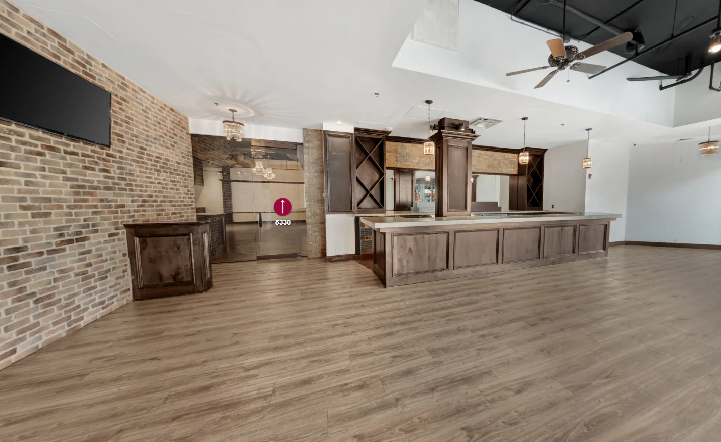 Spacious room with wood floors, brick accent wall, and a large wooden bar counter; pendant lights hang from the ceiling.
