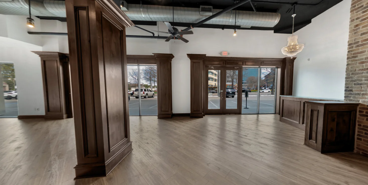 Spacious, empty room with wood floors, large windows, wooden columns, chandelier, and exposed ceiling ducts; view of parked cars outside.