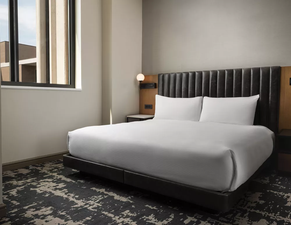 King bed with a gray leather bed frame and head board with white linen sheets a reading lamp attached to the wooden head board with an white and black night stand in the ADA Accessible Suite bedroom at Dallas, TX hotel