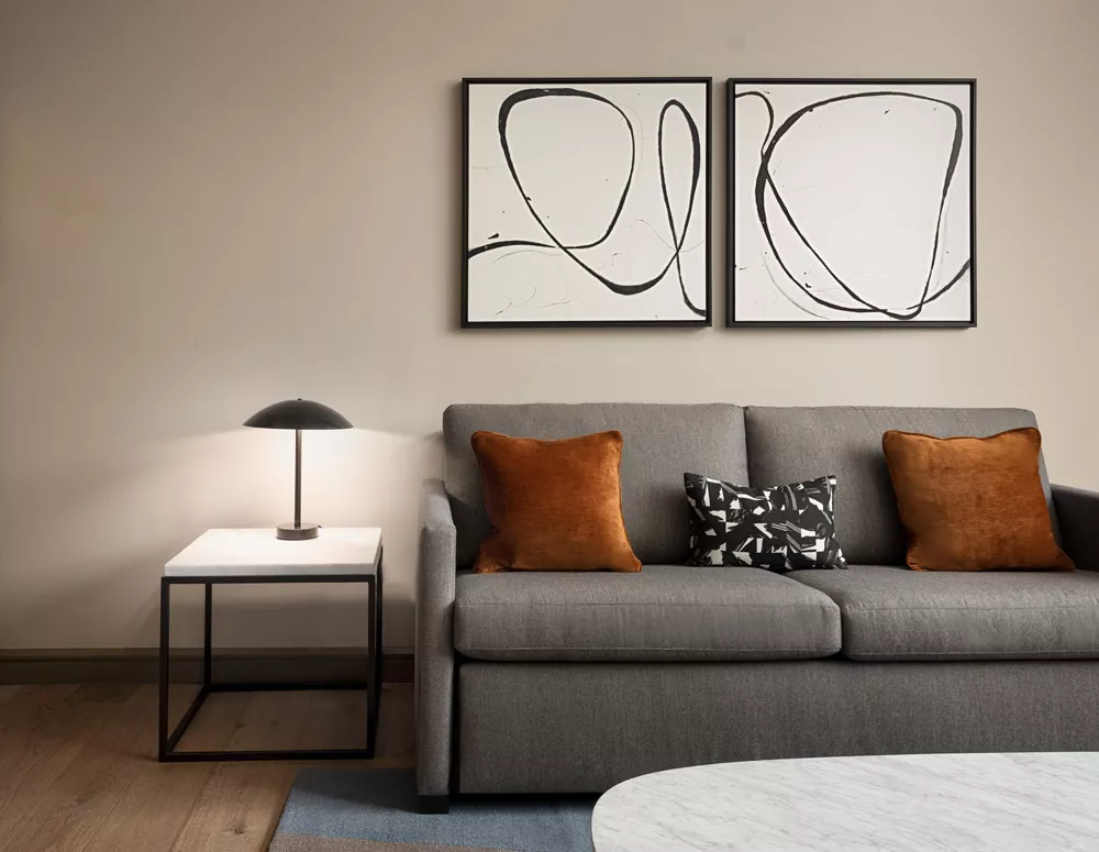 Gray couch with burnt orange and black pillows a white and black end table with a black reading lamp and black and white art on the wall in an ADA Accessible Suite in Dallas, TX at the Highland Dallas Curio Collection hotel
