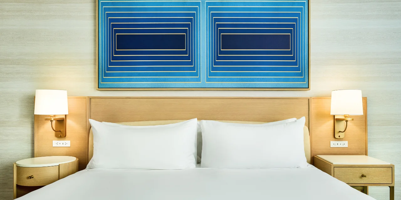 A neatly made hotel bed with crisp white linens, light wood furniture, and a large geometric blue artwork hanging above the headboard.