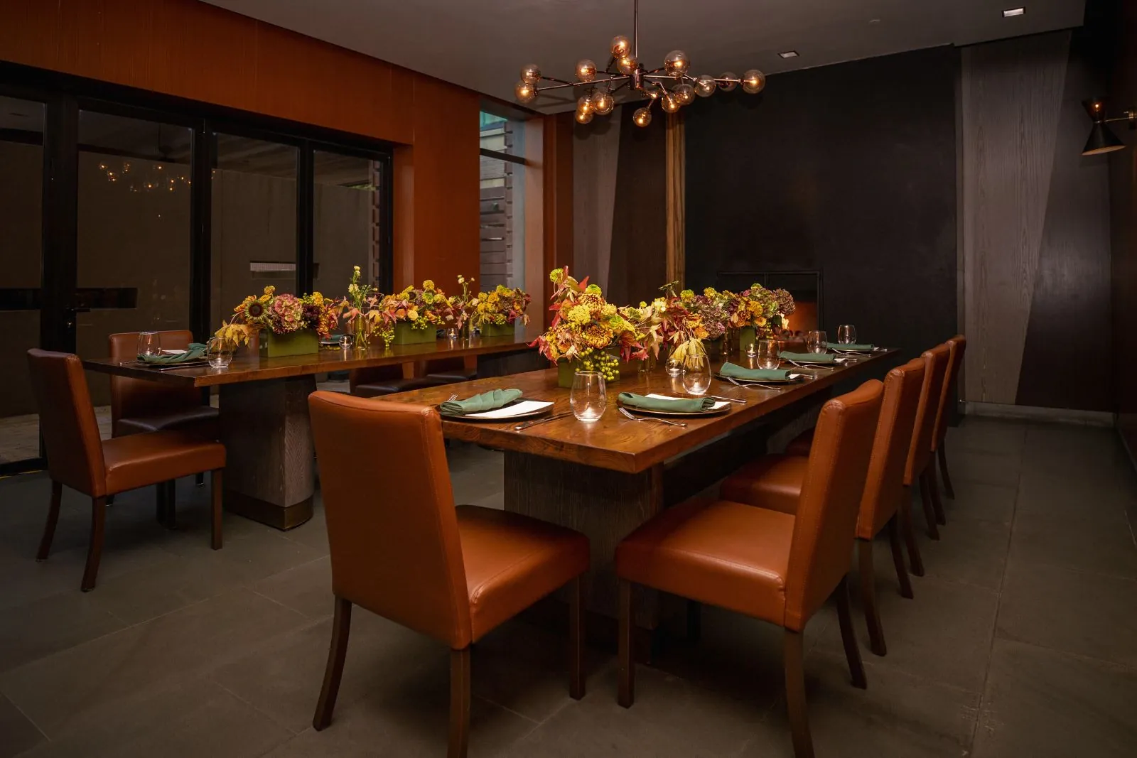 Private dining room with two long wooden tables, leather chairs, floral centerpieces, and warm modern lighting.