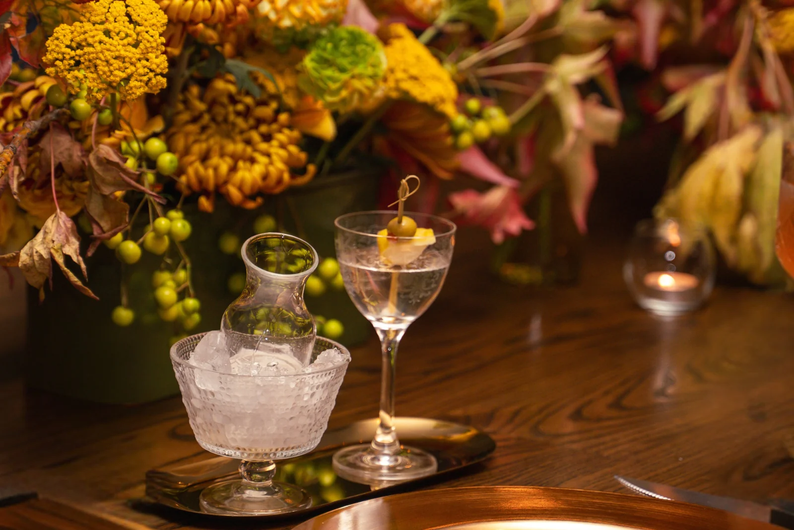 A chilled martini with an olive garnish and a small carafe set on ice, placed on a wooden table with autumn floral arrangements in the background.