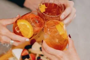 Three people clinking glasses of orange cocktails over a charcuterie board.