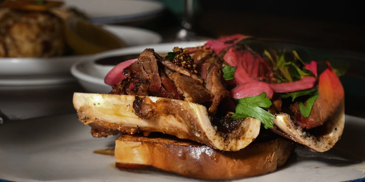 A gourmet dish featuring roasted bone marrow topped with tender sliced beef, pickled onions, fresh herbs, and mustard seeds, served on toasted bread.