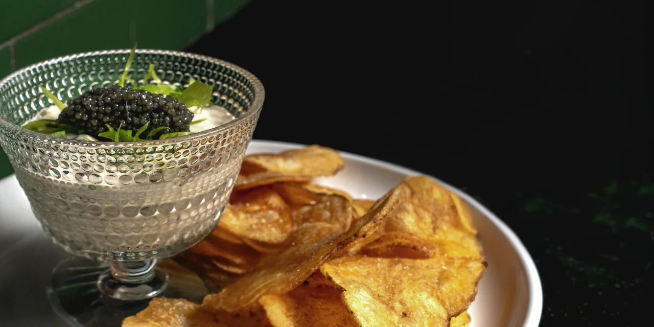A glass bowl filled with creamy dip topped with black caviar and fresh greens, served alongside a plate of golden, crispy potato chips.