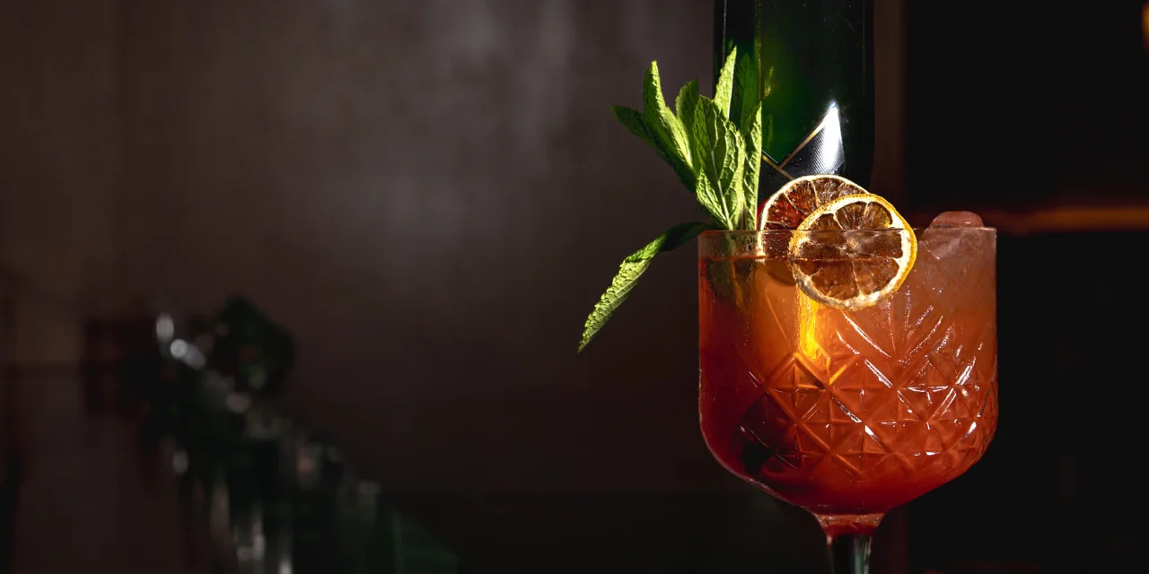 A ruby-red cocktail served in a cut-crystal glass, garnished with fresh mint leaves and dried citrus slices, set against a dark, moody bar backdrop.