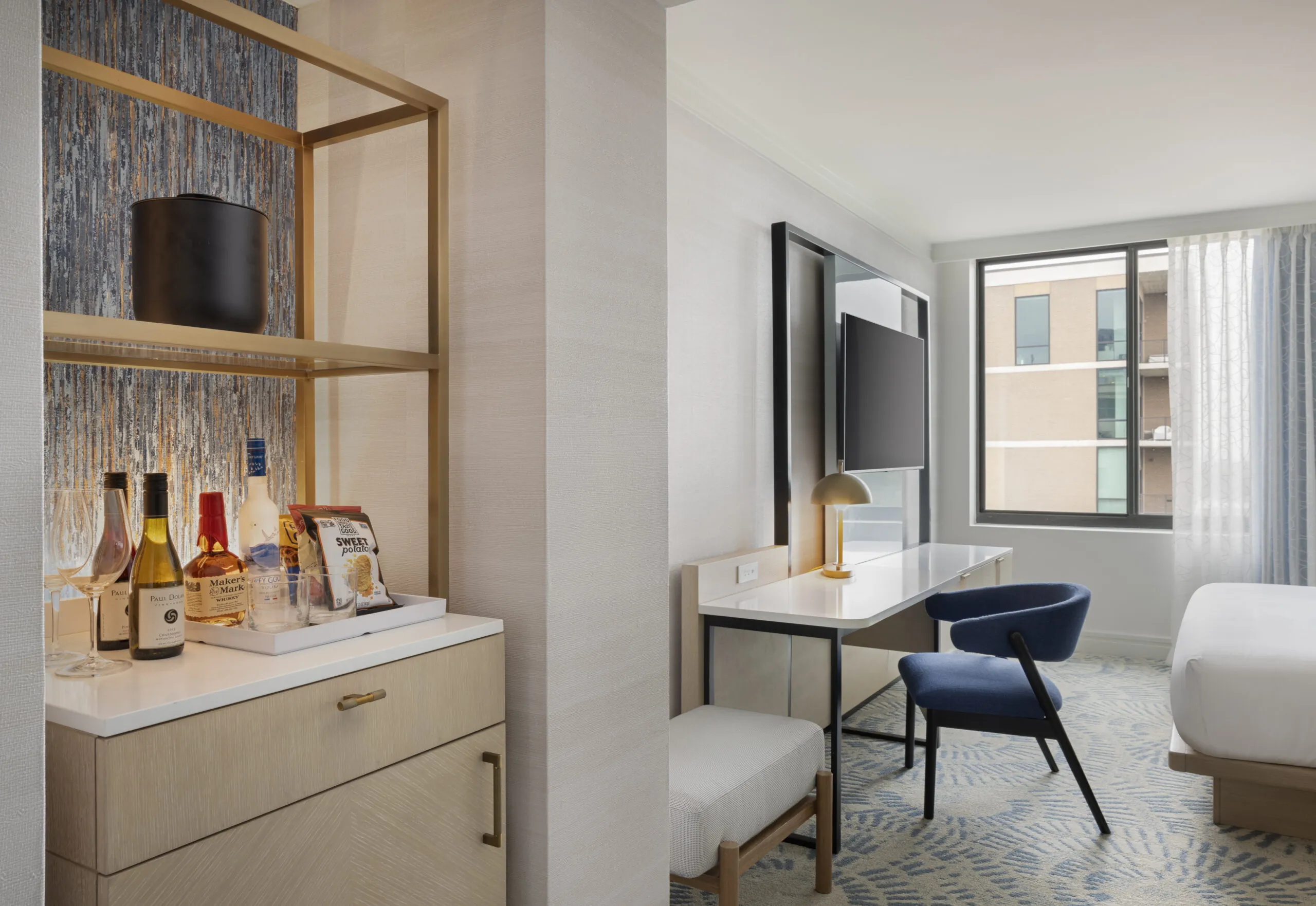 Hotel room with a minibar stocked with drinks and snacks, a desk with a chair and lamp, TV on the wall, bed, and a large window with curtains.