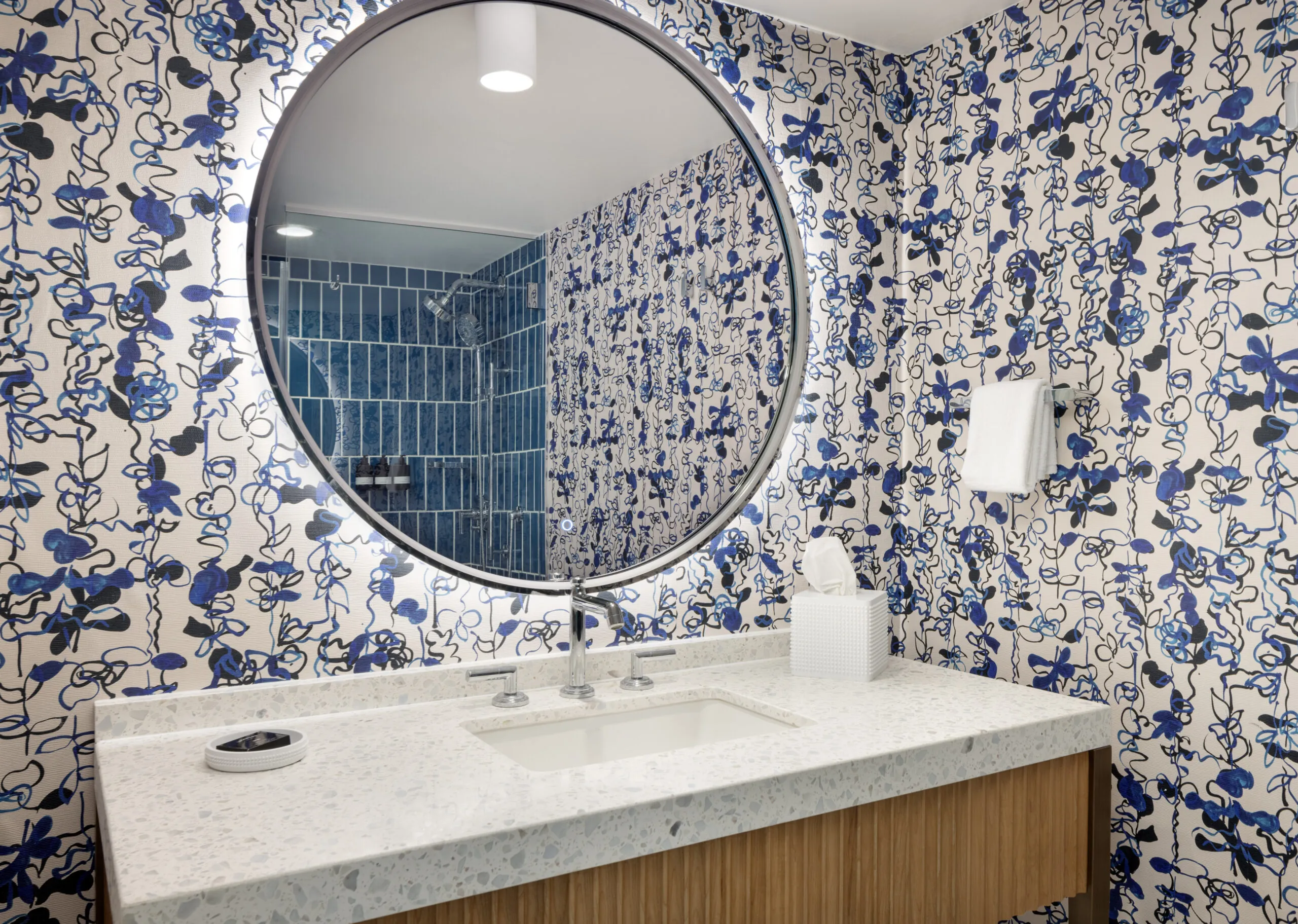 A bathroom vanity with a round mirror, blue floral wallpaper, a white sink, tissue box, and blue-tiled shower reflected in the mirror.