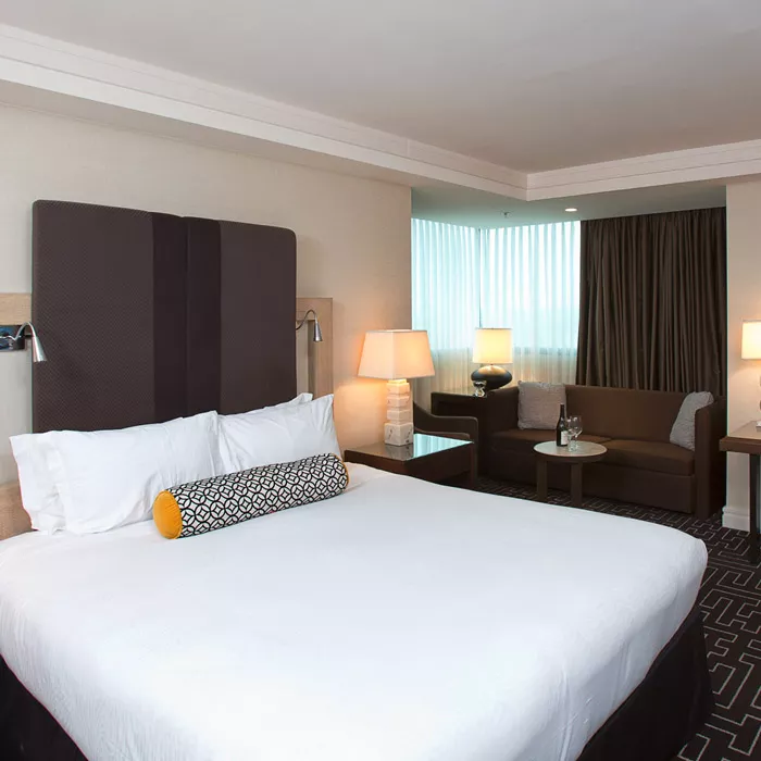 King guest room at Highland Dallas, TX with white linen sheets and a brown head board with reading lamps, brown couch with a small circular coffee table with a bottle of wine and wine glass on it, a coffee table with a white lamp, corner floor to ceiling windows with brown curtains