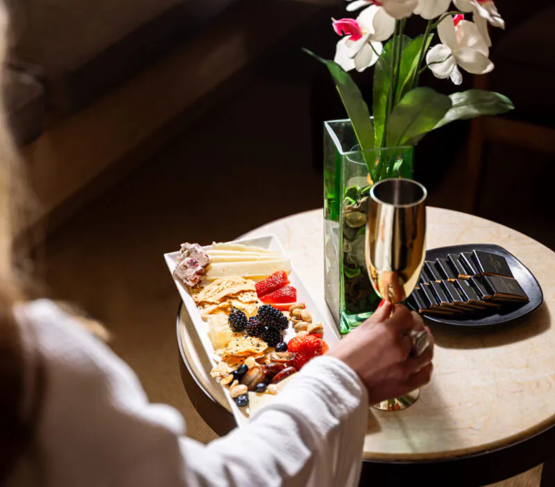 A person holding a champagne glass sits by a table with a cheese and fruit platter, chocolate, and a vase with white and pink flowers.
