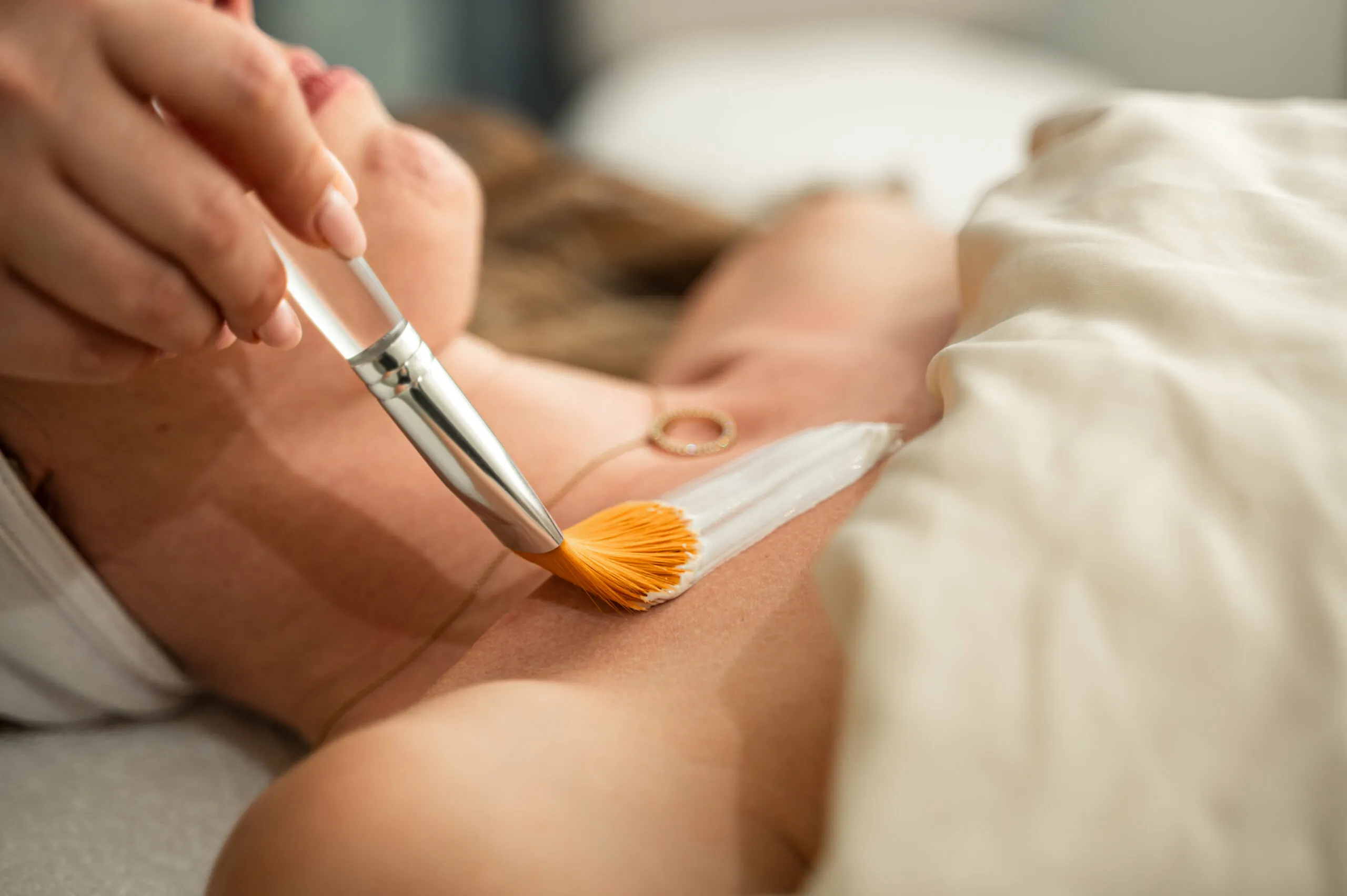 A close-up of a spa treatment in progress, showing a brush applying a cream or mask to a woman’s chest as she relaxes under a soft blanket.