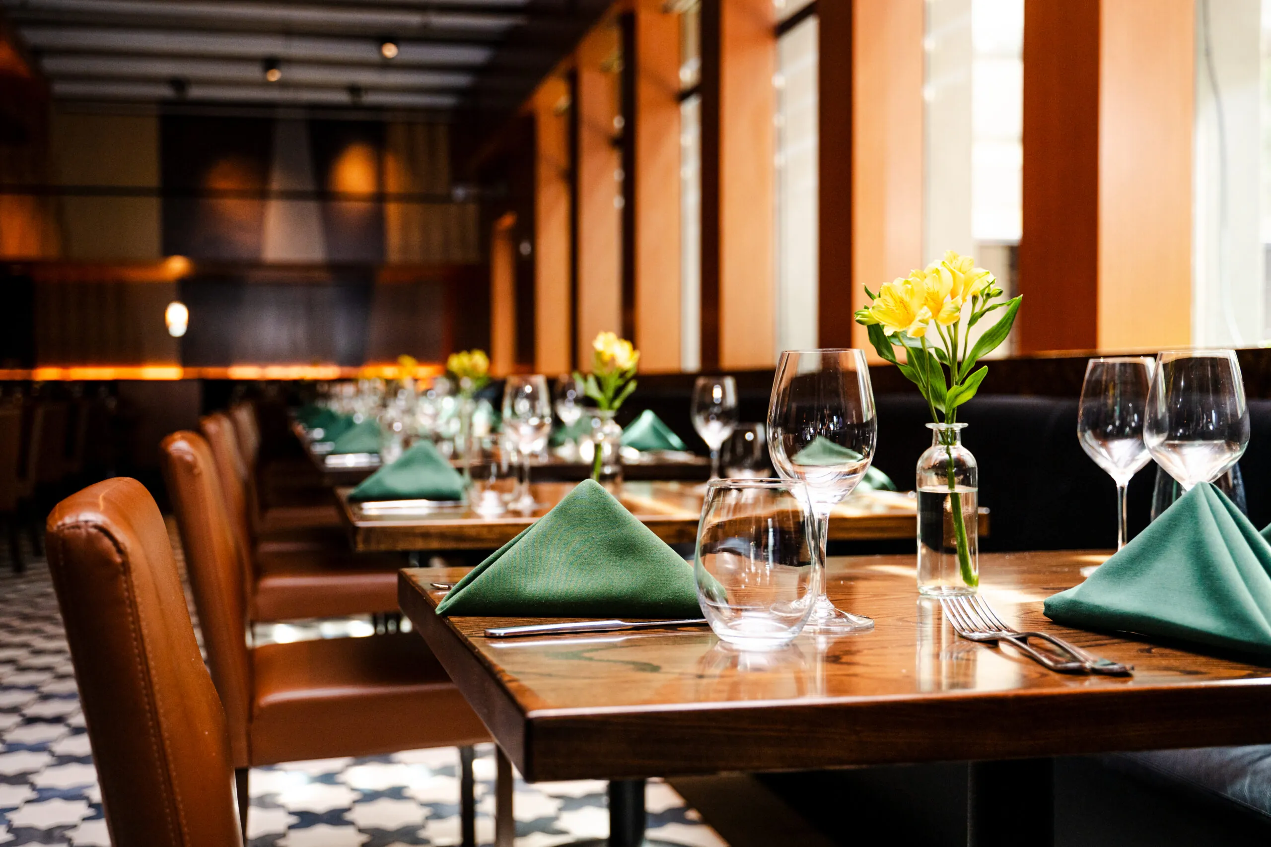 Elegant dining room at The Reserve with wooden tables set with wine glasses, folded green napkins, and small yellow flower arrangements in glass vases, creating a warm and inviting atmosphere.