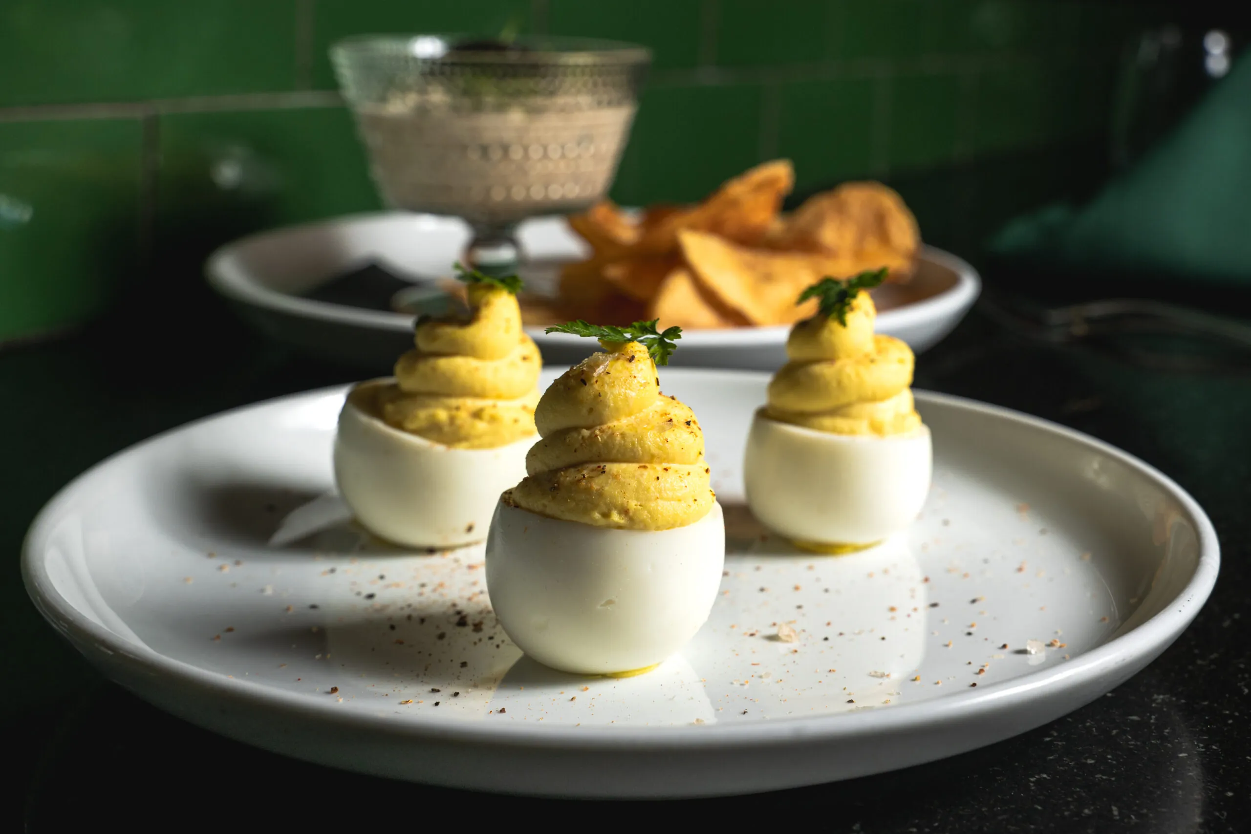 A trio of deviled eggs with creamy, piped yolk filling and a garnish of fresh herbs, served on a white plate at The Reserve. A bowl of house-made chips and dip is blurred in the background.