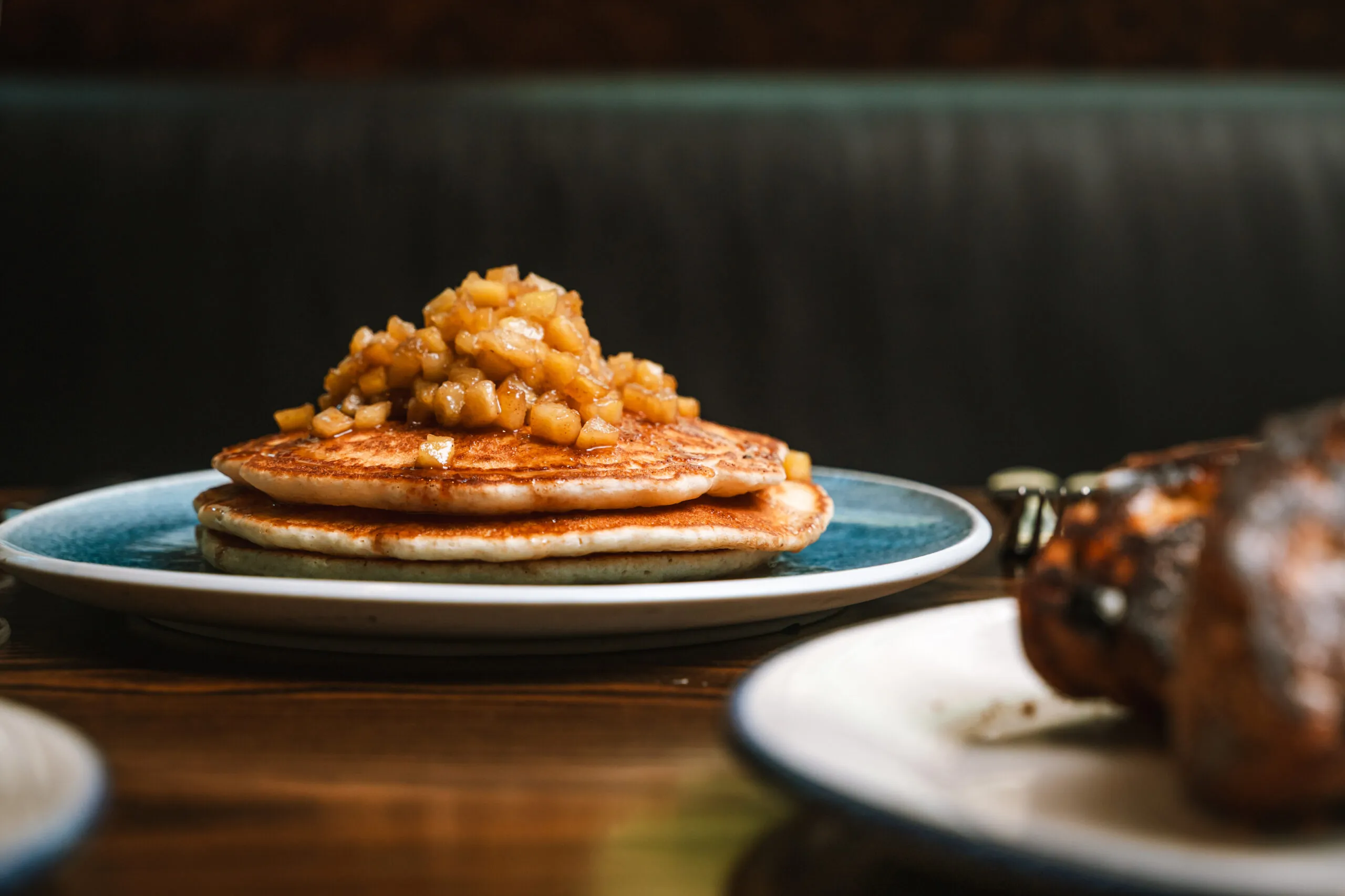 A stack of pancakes topped with diced caramelized apples sits on a blue and white plate, placed on a wooden table.