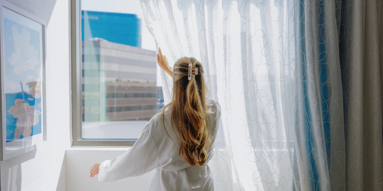 A woman in a white robe pulls back sheer curtains to reveal a view of modern city buildings under bright daylight.