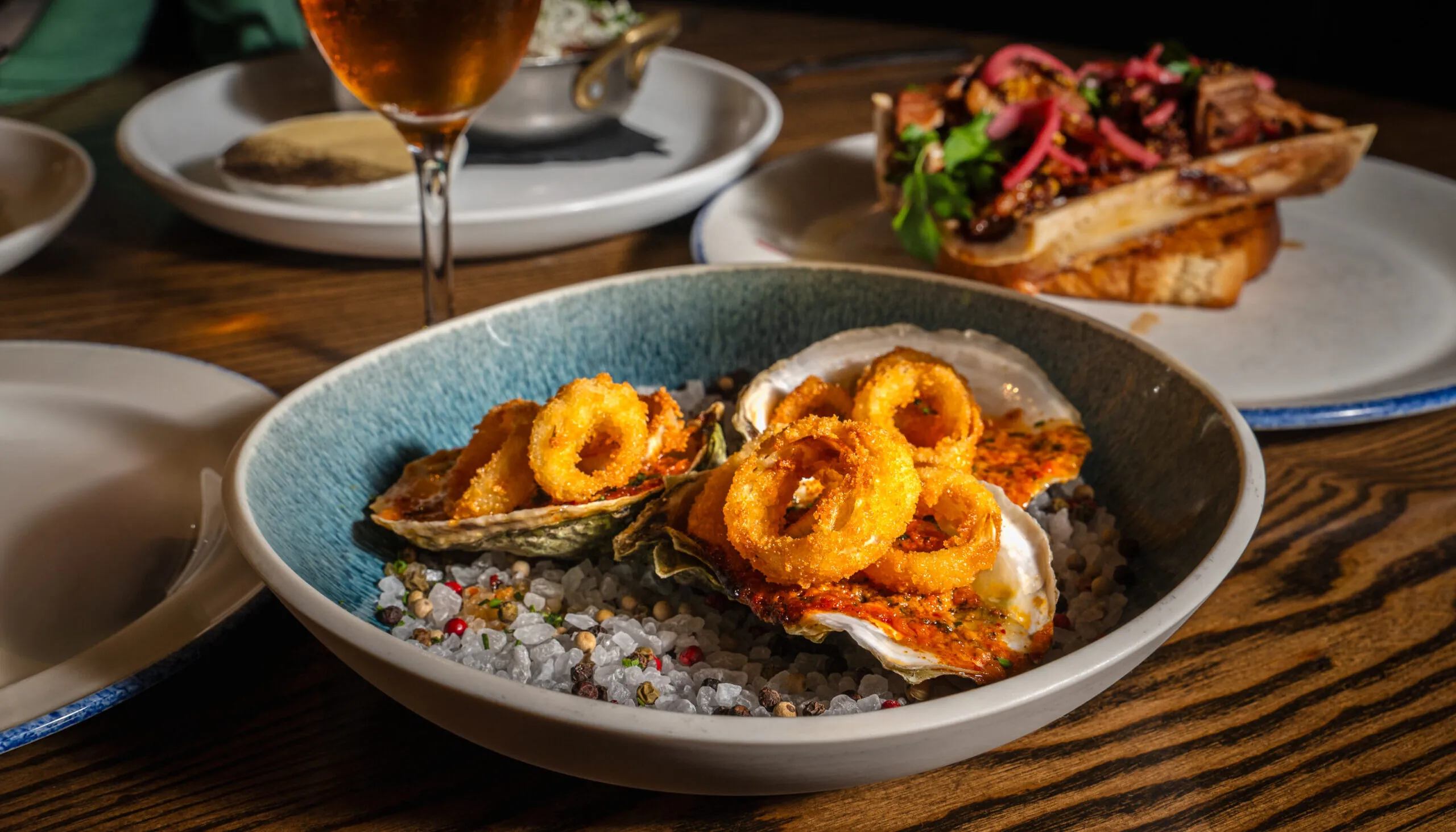 A bowl of grilled oysters topped with golden fried calamari rings, served on a bed of coarse salt and peppercorns. The dish is paired with a cocktail garnished with a black cherry and surrounded by other plates, including bone marrow toast, on a wooden table in a dimly lit restaurant setting.