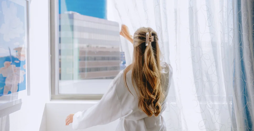 A woman with long blonde hair, wearing a white robe, opens sheer curtains to reveal a city view from her hotel room on a bright morning.