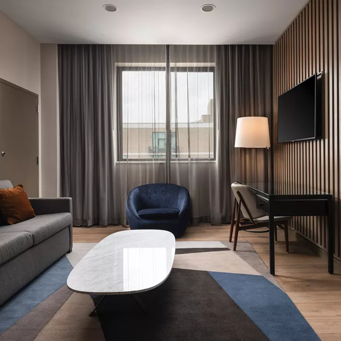 Accessible suite at Highland Dallas with a gray lounge couch a white marble coffee table, a wooden desk area with a white and brown desk chair and a flat screen tv on the wall with a window with gray drapes at the Highland Dallas, TX hotel