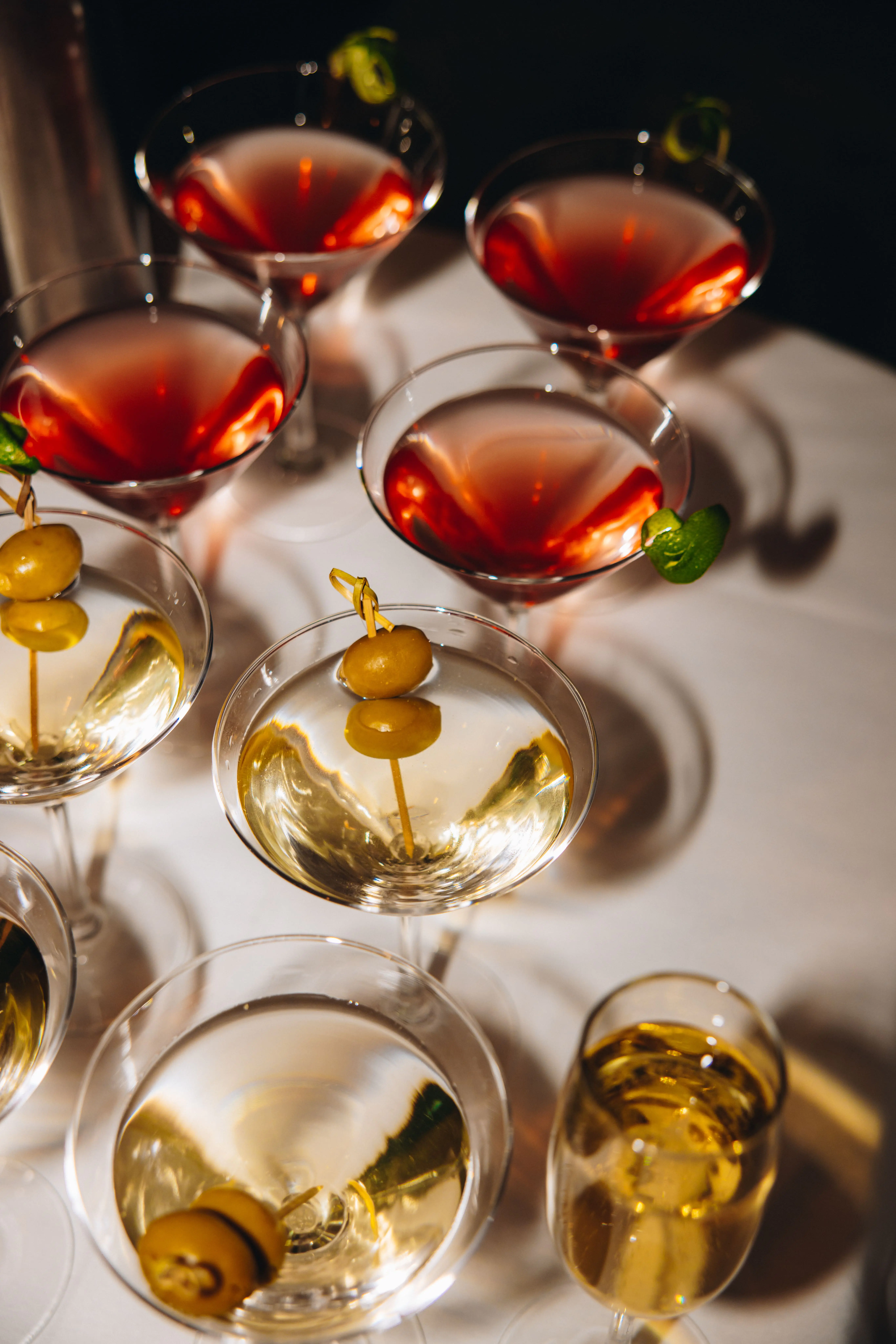 Seven martini glasses with olives or lime garnishes and one tumbler glass with a yellow drink, arranged on a white tablecloth in bright light.