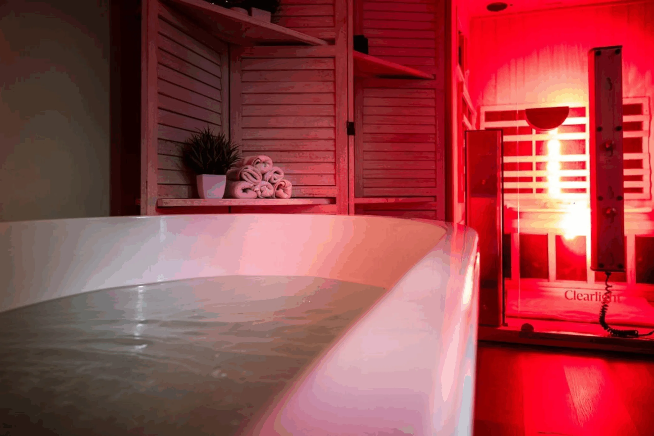 Freestanding soaking tub filled with water inside a softly lit spa suite, with folded towels on a shelf and an illuminated infrared sauna glowing red in the background.