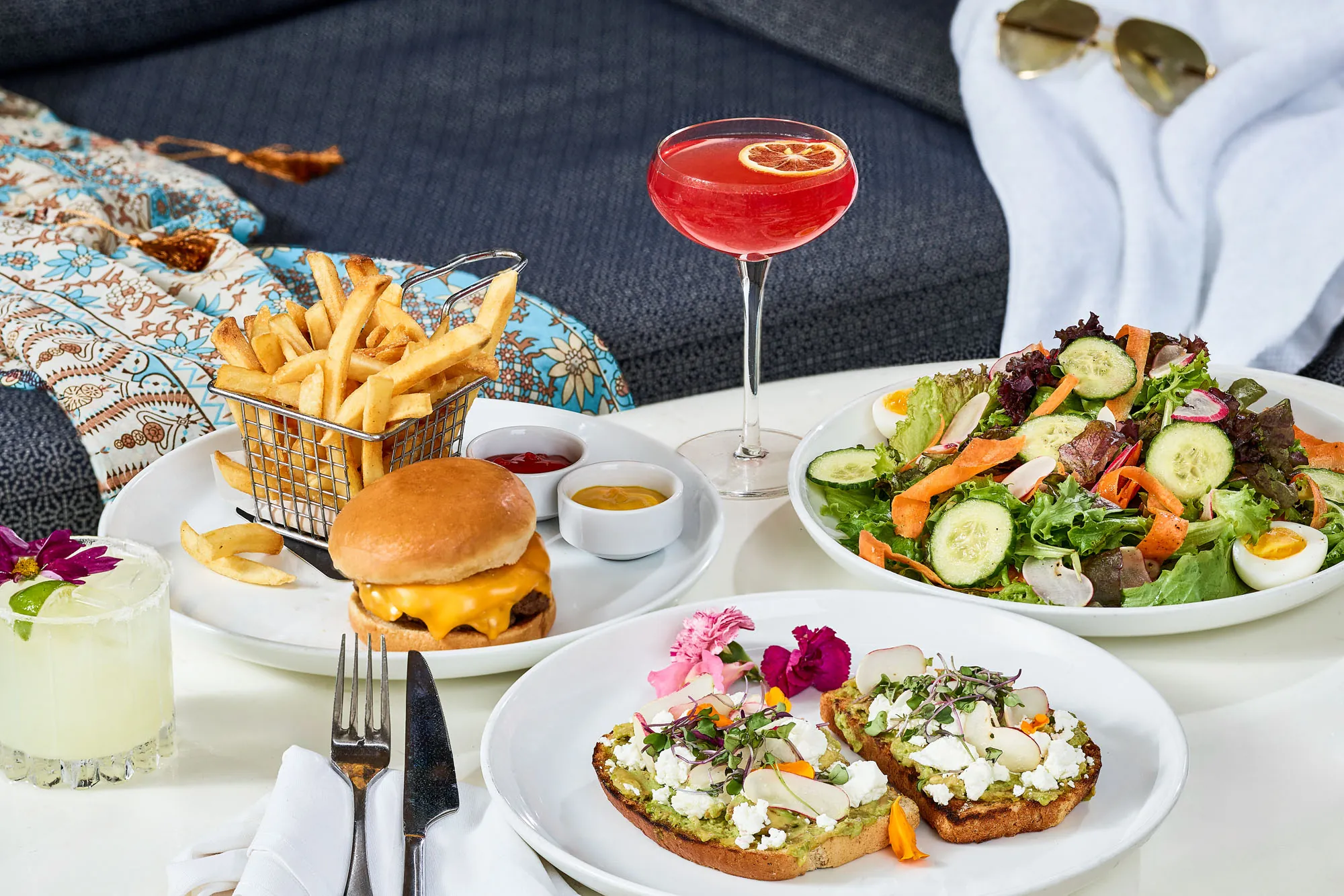 A table with a cheeseburger and fries, two cocktails, a fresh salad, and two open-faced avocado toasts, set by a patterned towel and sunglasses.