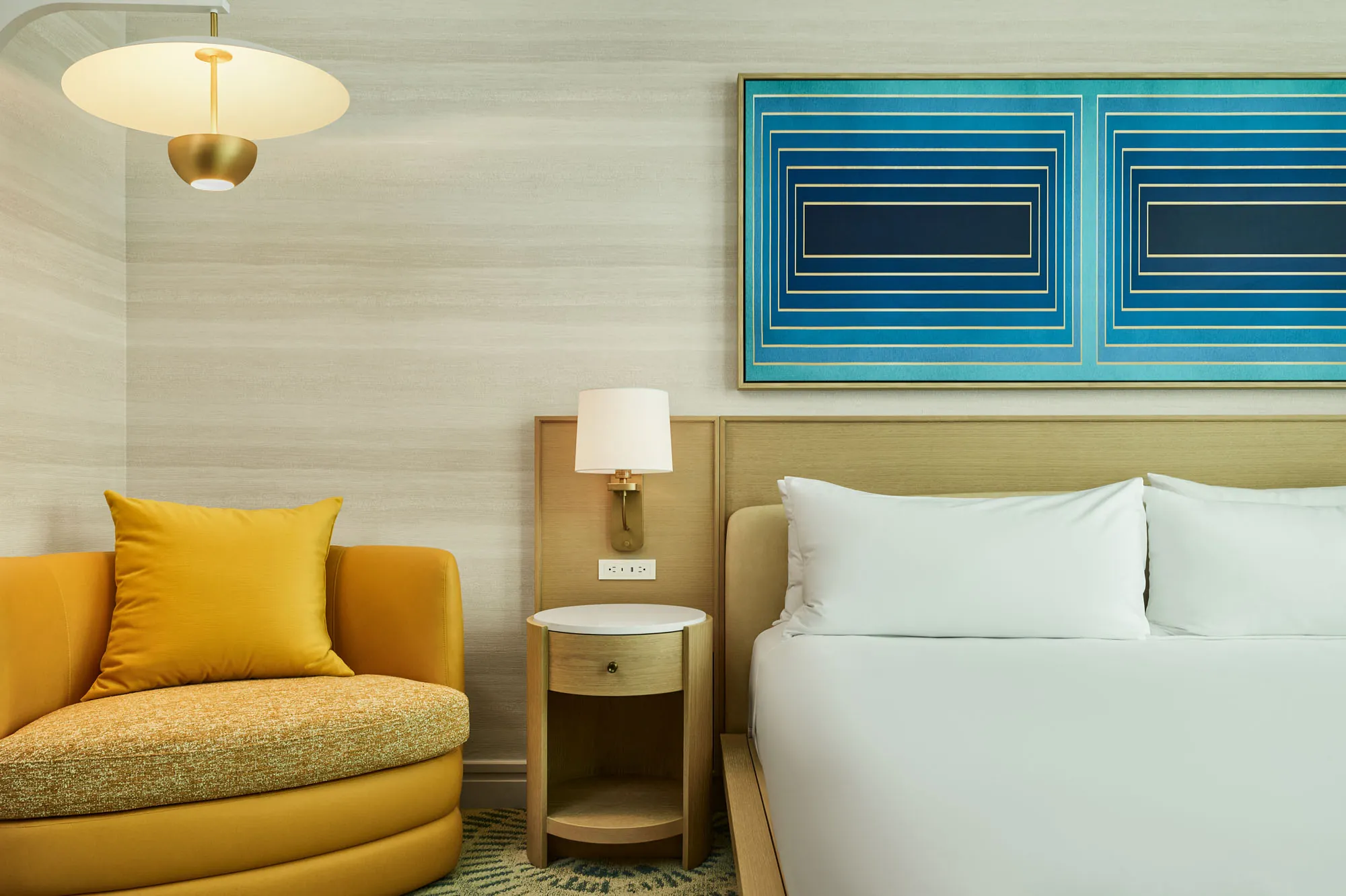 Hotel guest room detail showing part of a neatly made bed with white linens, a light wood nightstand and wall sconce, a mustard armchair with pillow, and bold geometric blue artwork above the headboard.