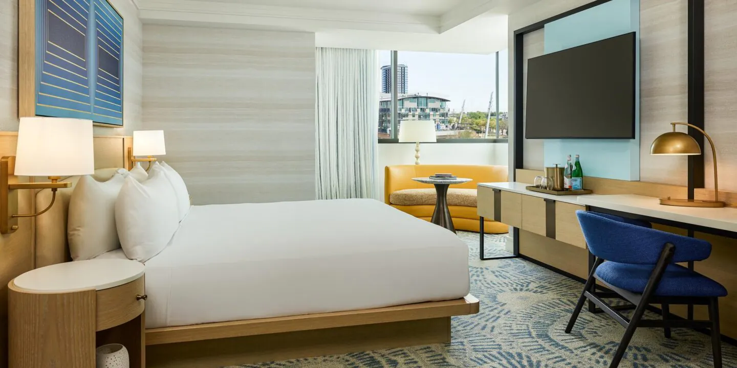 Modern hotel room with a king bed, desk, TV, lounge chair, large window, and neutral decor with blue accents.