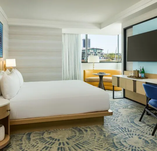 Modern hotel room with a king bed, desk, TV, lounge chair, large window, and neutral decor with blue accents.