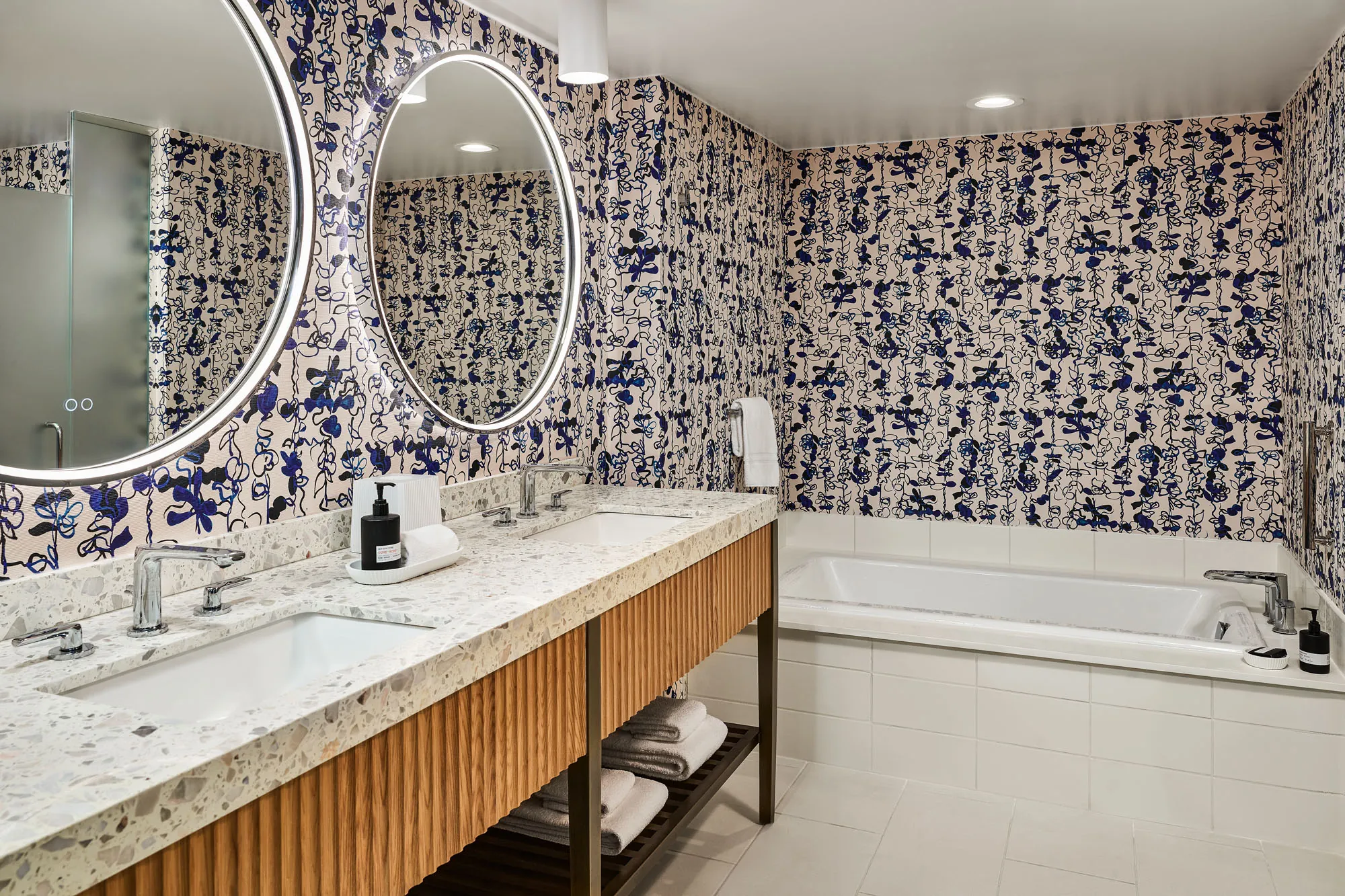 Modern bathroom with two sinks, large round mirrors, patterned wallpaper, a bathtub, and neatly folded towels on shelves under the counter.