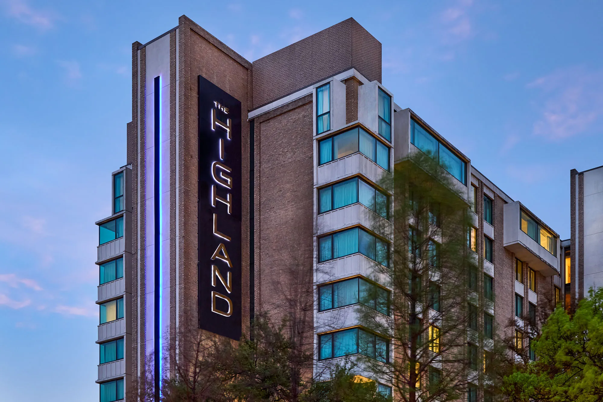 Close-up view of The Highland Dallas hotel exterior at dusk, highlighting the tall illuminated vertical “Highland” sign and modern glass windows framed by brick architecture.