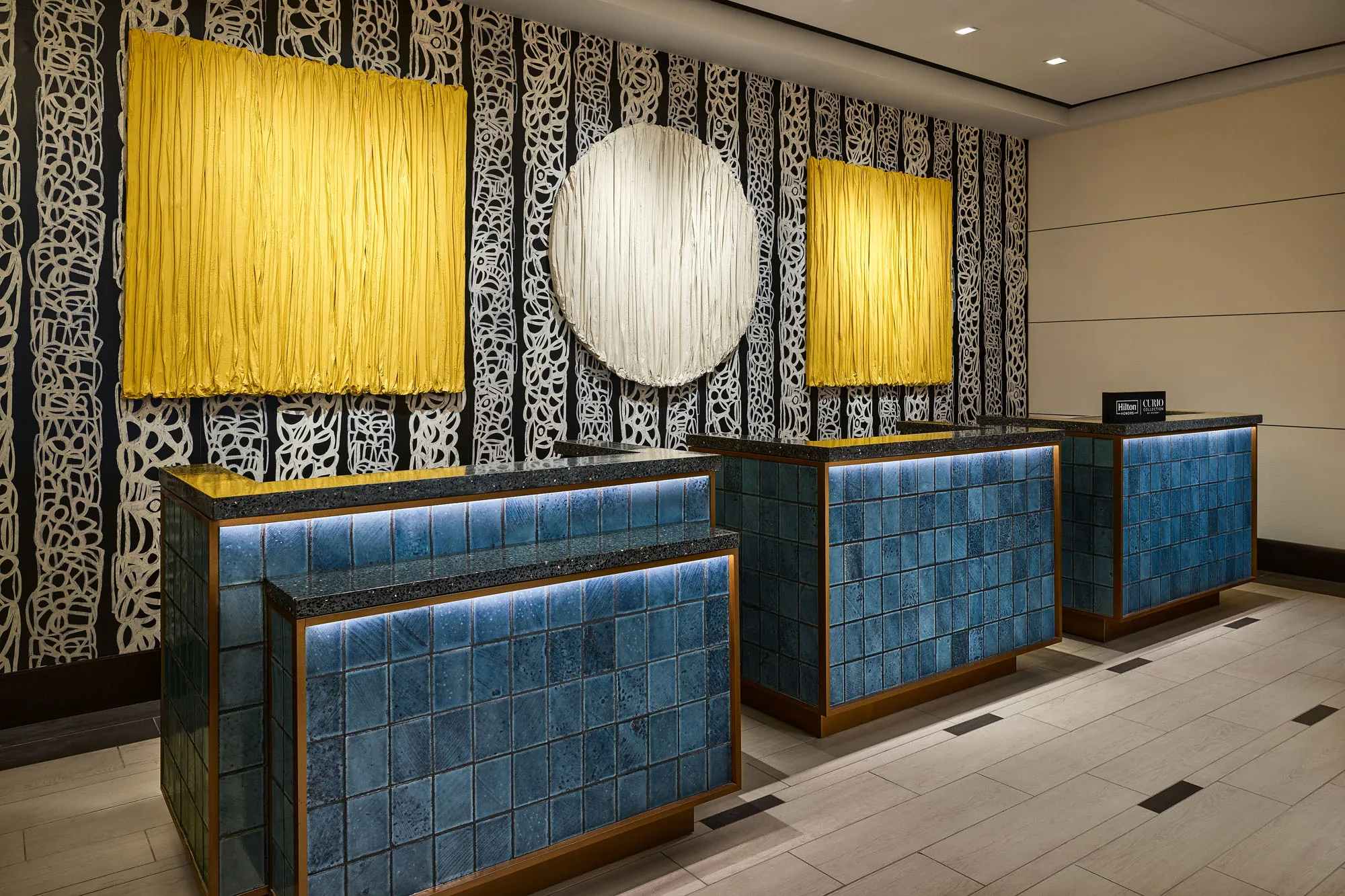 Hotel front desk area with three check-in counters clad in blue tile and accented by underlighting, set against a bold patterned wall featuring two large yellow textile art pieces and a central round white artwork.