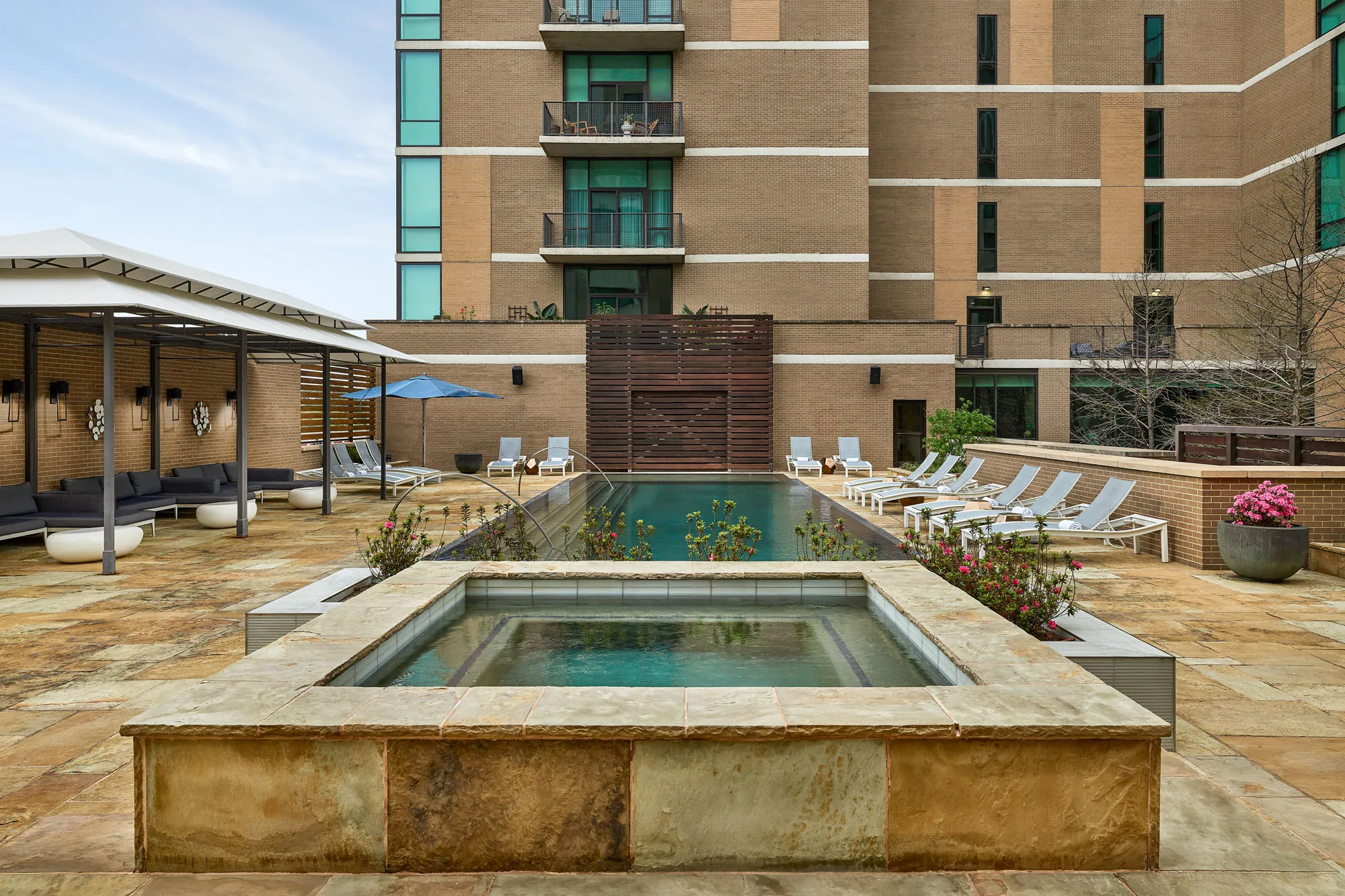Modern outdoor pool area with lounge chairs, a small hot tub in front, shaded seating to the left, and a tall apartment building in the background.