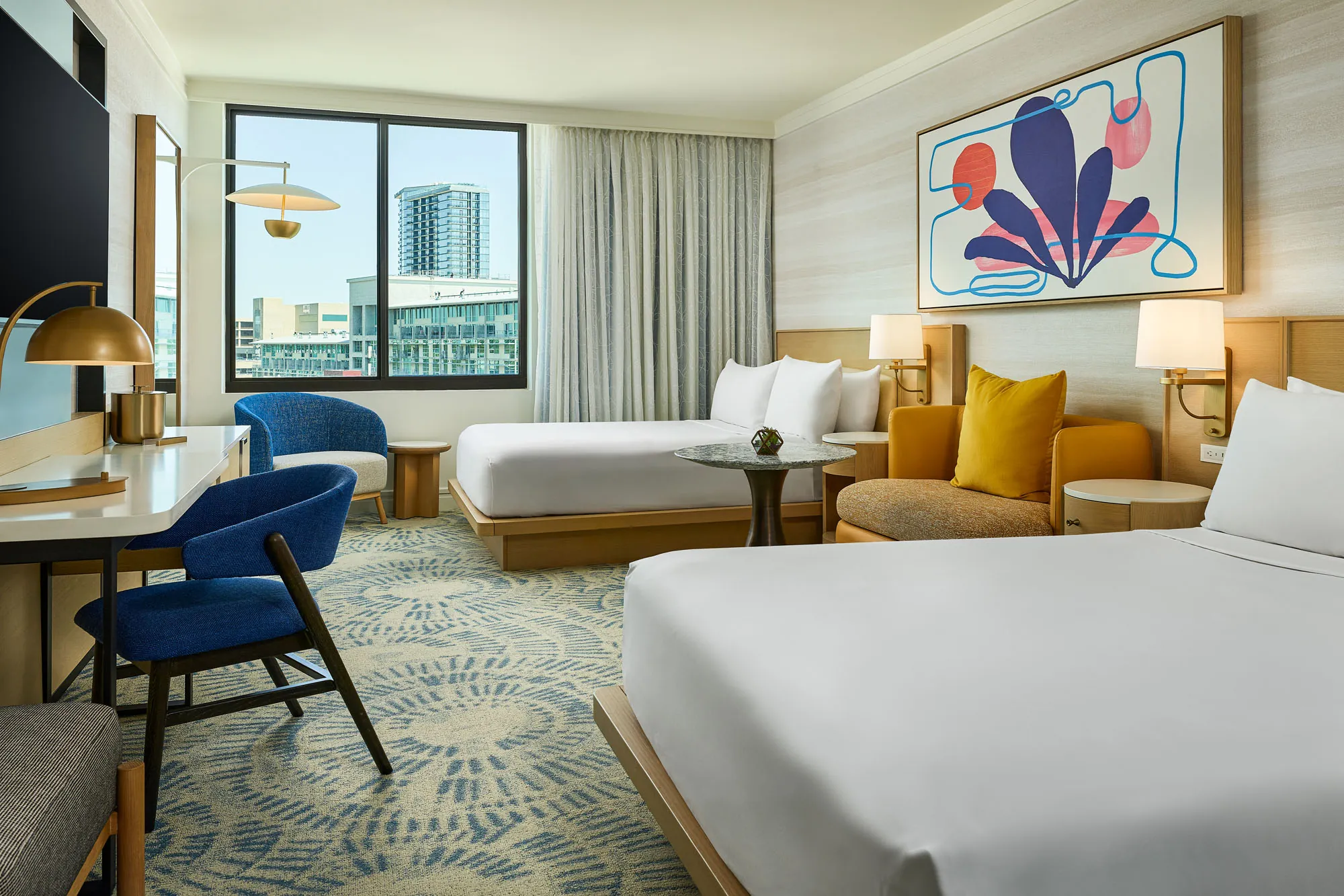 Queen Queen Deluxe room featuring two neatly made queen beds with white linens, modern wood furnishings, a work desk with blue chairs, cozy seating with mustard accents, abstract artwork, and large windows offering city views.