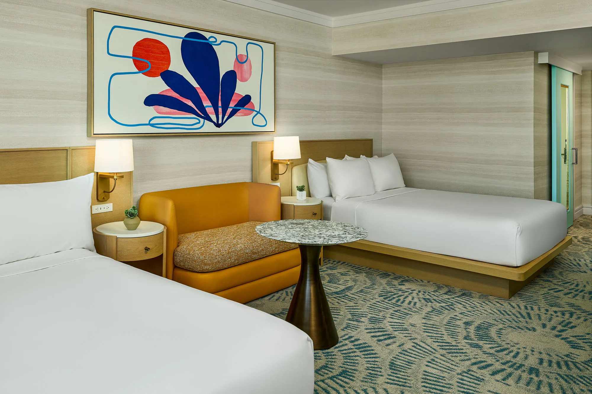 Queen Queen Deluxe room with two queen beds dressed in crisp white linens, a mustard loveseat with patterned cushion, round stone-top table, modern bedside lighting, and abstract artwork above the beds.