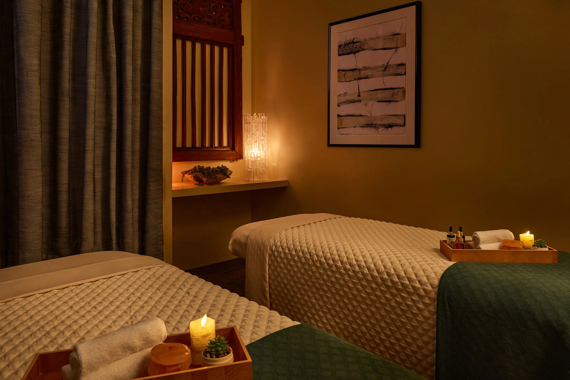 Couples spa treatment room with two massage tables dressed in textured linens, softly lit by candles and warm ambient lighting, featuring neatly arranged trays with rolled towels, oils, and decor for a calming atmosphere.