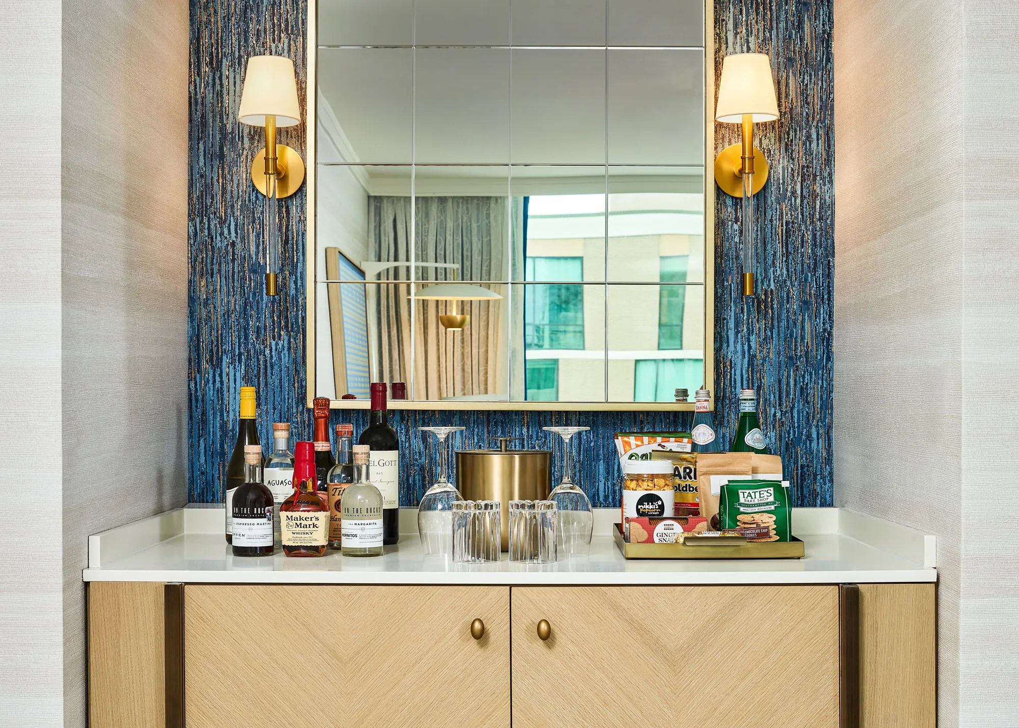 A minibar setup with liquor bottles, snacks, glasses, an ice bucket, and a large mirror, flanked by two wall sconces with gold accents.