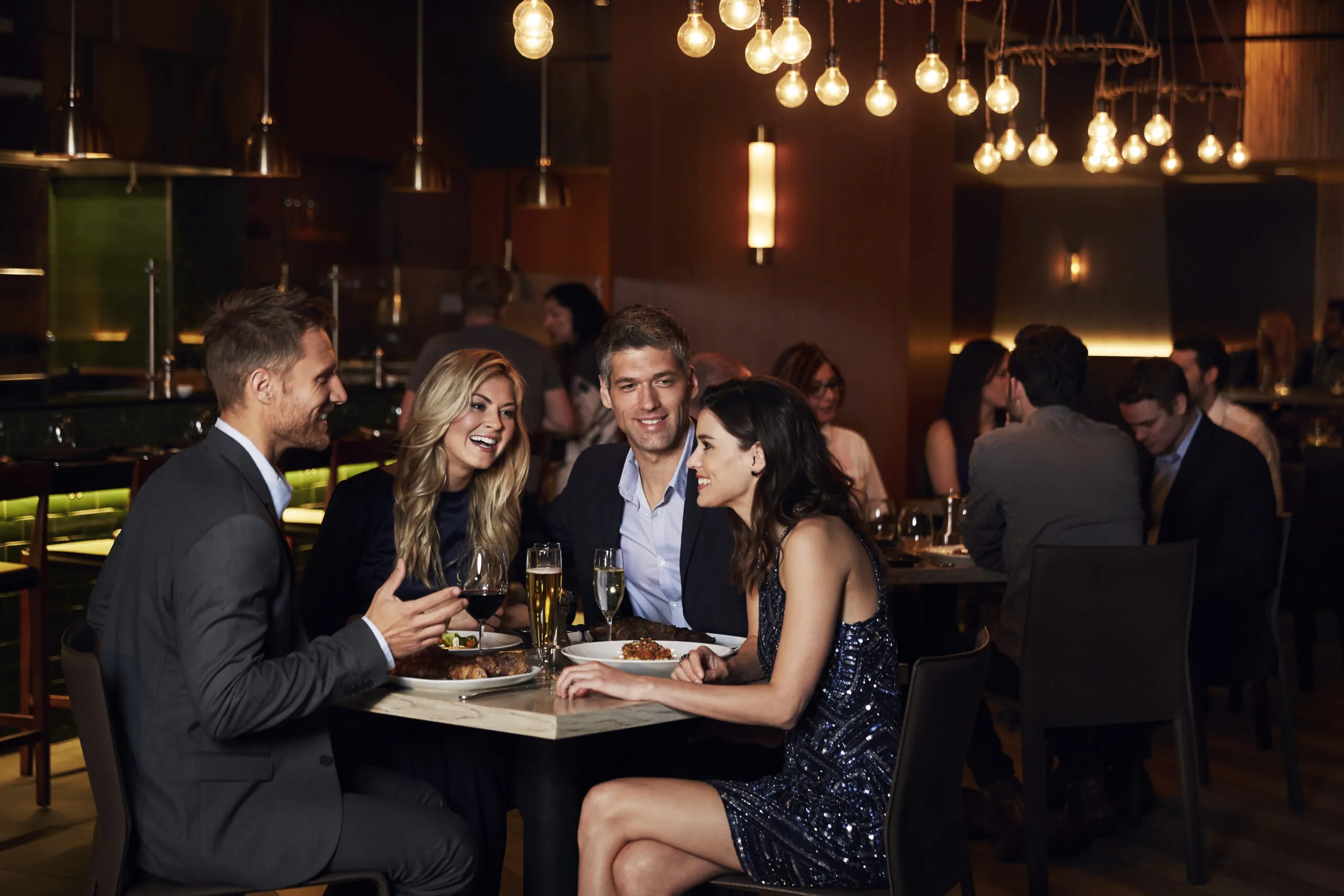 Four people dressed up, sitting at a restaurant table, talking and laughing, with drinks and food in front of them, and other diners in the background.