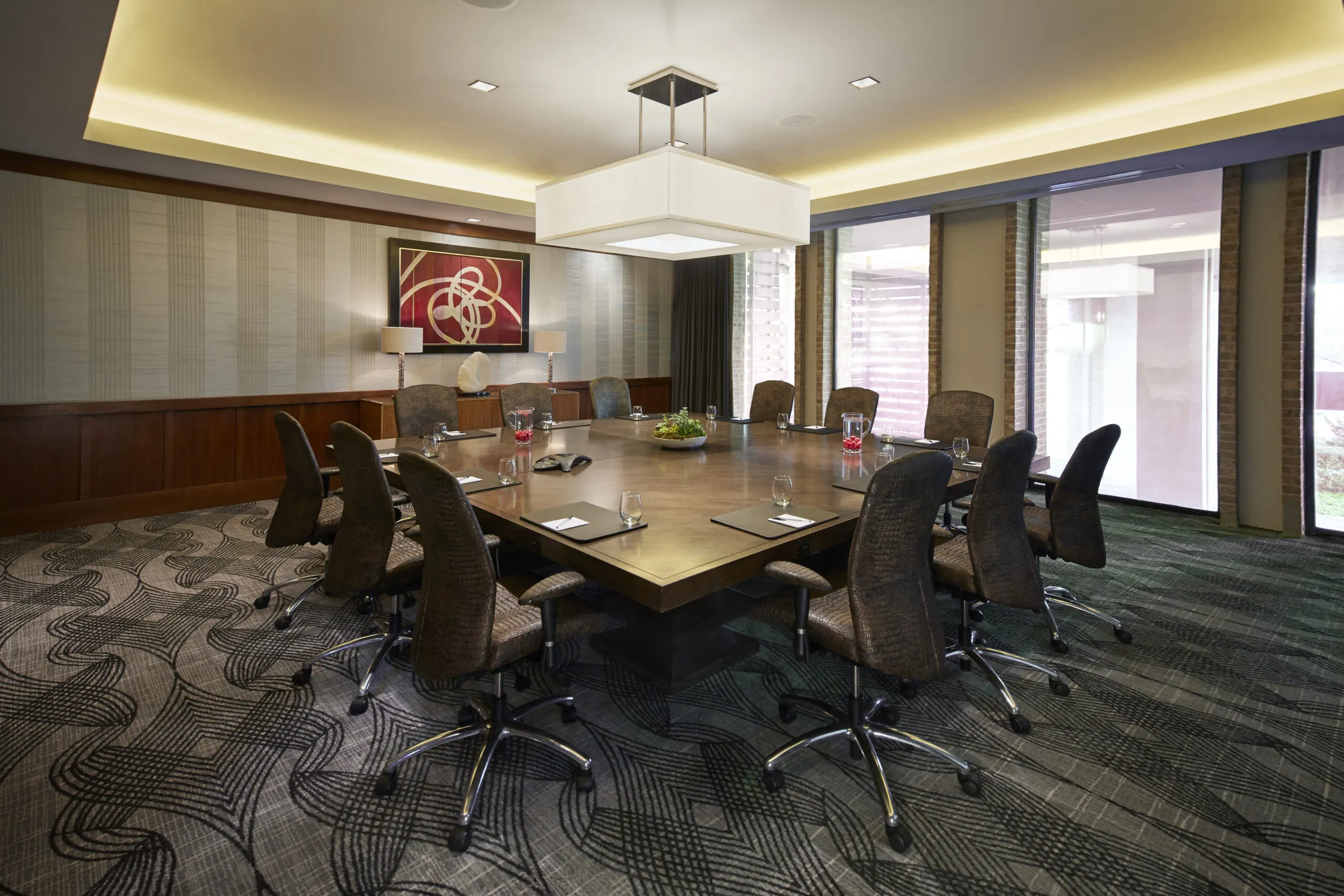 Modern conference room with a large square table, ten chairs, water glasses, and a painting on the wall under soft ceiling lights.