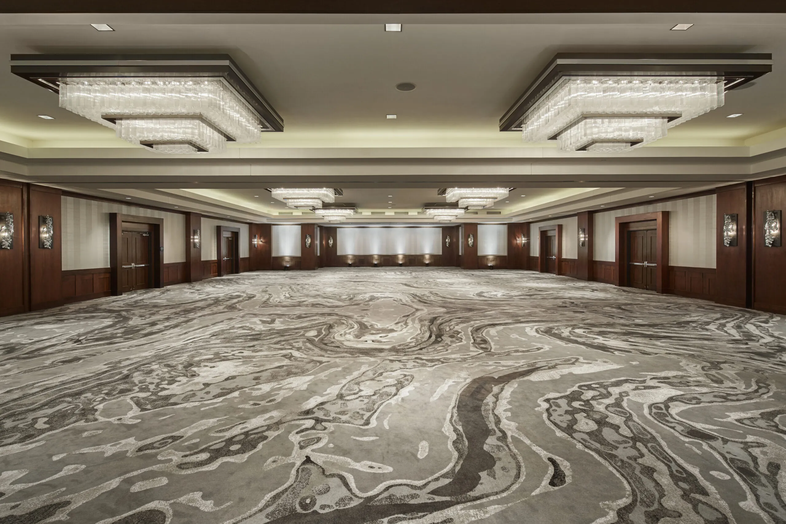 Spacious, empty hotel ballroom with modern chandeliers, patterned carpet, wood-paneled walls, and closed double doors on each side.
