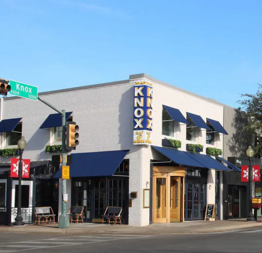 A white corner building with blue awnings, large windows, and a vertical “KNOX” sign; street signs read “Knox” at an intersection.