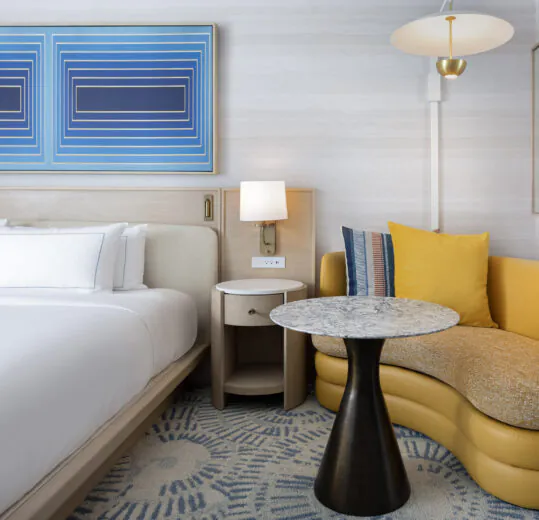 Modern hotel room with a white bed, yellow sofa, round marble-top table, wall art, and lamps on a light-patterned carpet.