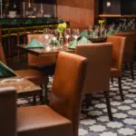 A cozy restaurant with brown chairs, set tables, green napkins, glassware, and yellow flowers in vases on a patterned tile floor.