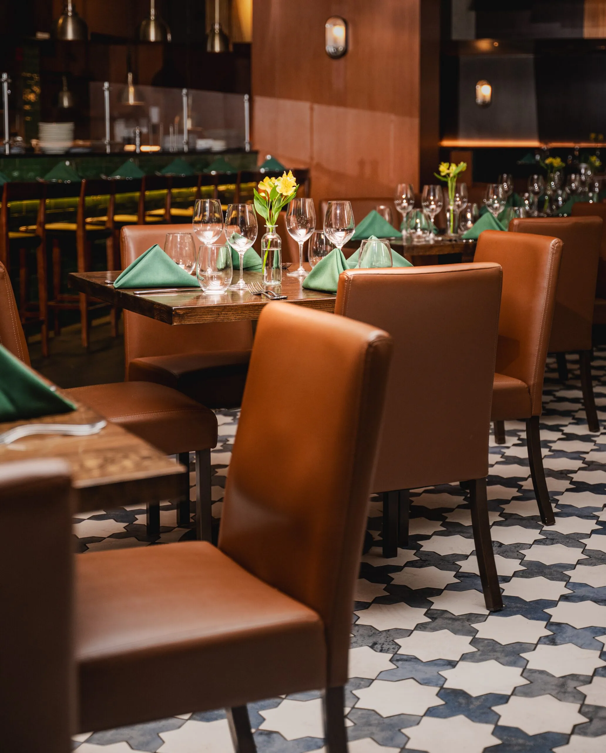 A cozy restaurant with brown chairs, set tables, green napkins, glassware, and yellow flowers in vases on a patterned tile floor.