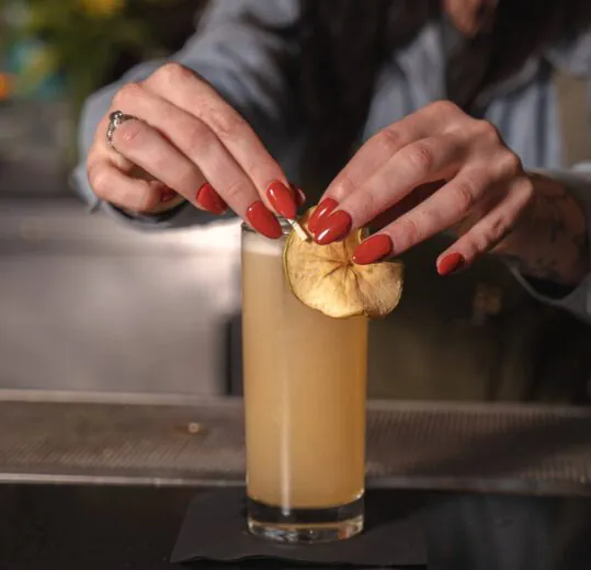 A person with red nails places a dried apple slice garnish on a tall glass of light brown cocktail at a bar.
