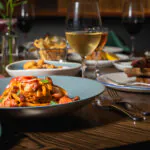 A plate of pasta with seafood and vegetables sits on a wooden table, surrounded by wine glasses and various dishes in a restaurant setting.